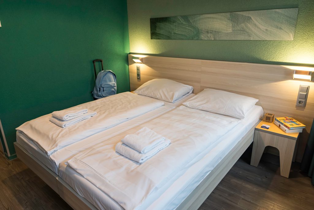 Helles Zweibettzimmer mit grüner Wand, weichen Betten und Rucksack