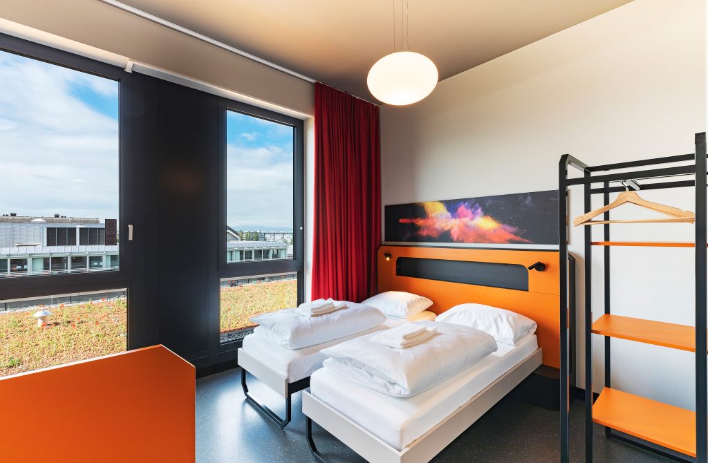 Bright twin room with orange accents, large windows, and city view