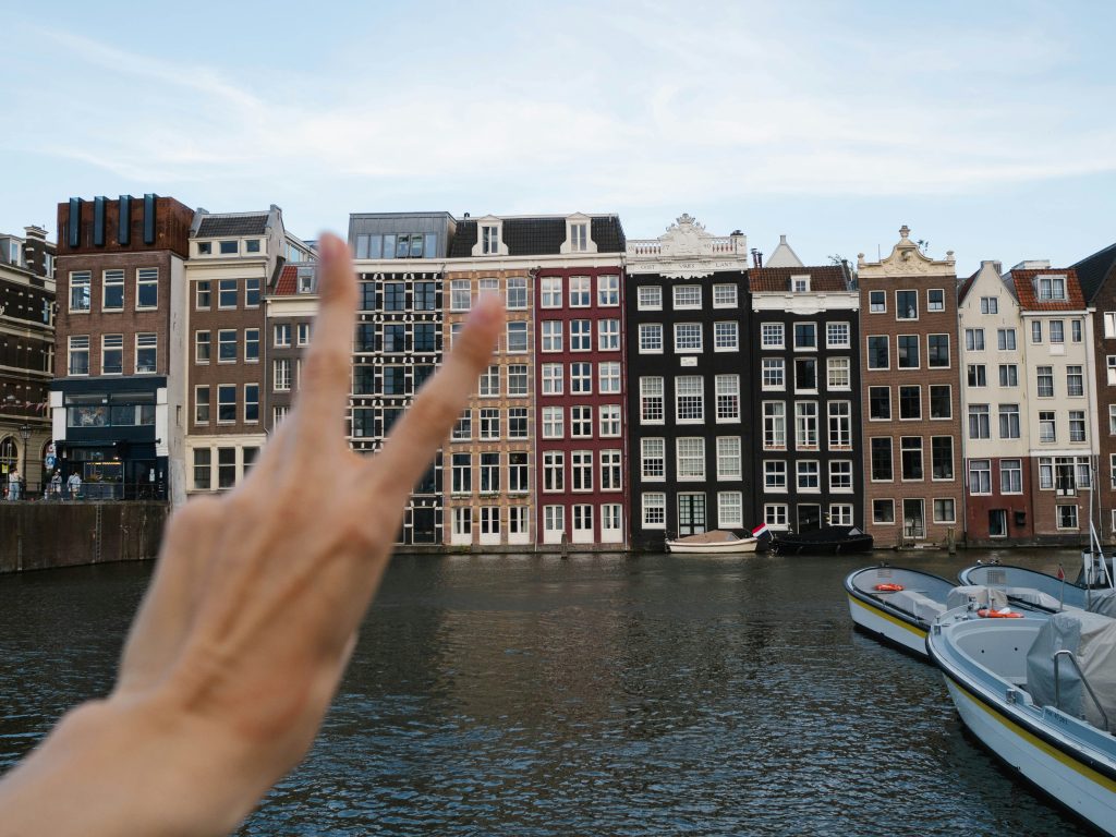 Hen party Amsterdam: hand showing a peace sign with Amsterdam buildings and canal on the background
