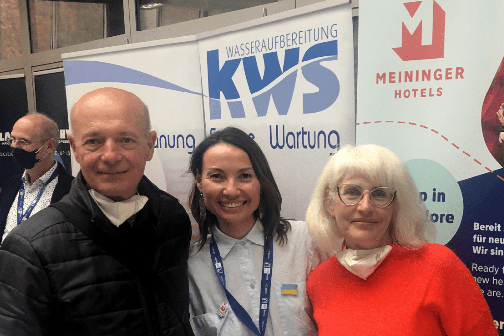 Zhanar hugging a man on the left and woman on the right with a job fair sponsors on the background