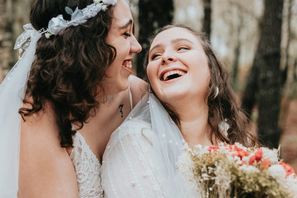 Heiraten in Kopenhagen: Zwei Bräute lachen in Hochzeitskleidern mit Blumenstrauß im Wald  