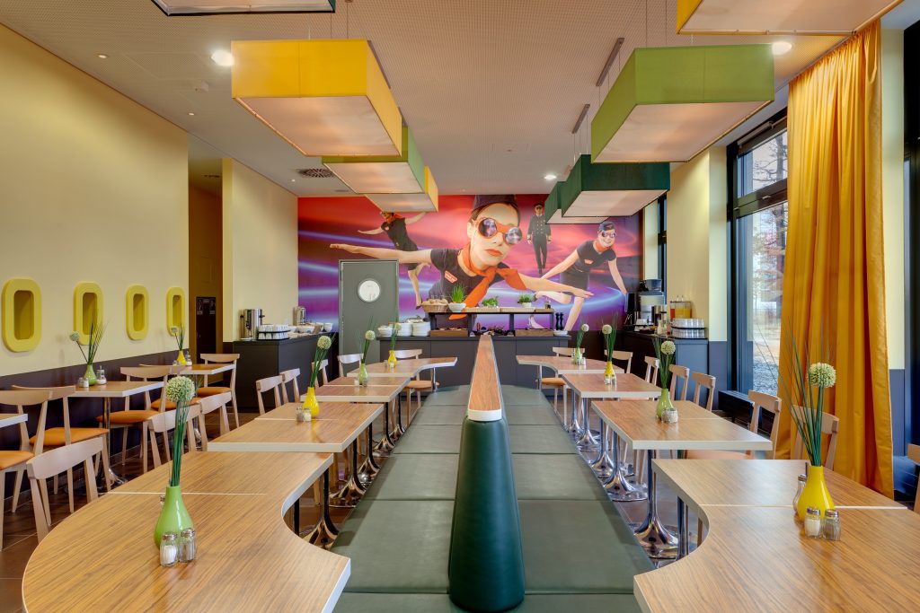 MEININGER Hotel Frankfurt/Main Airport: sala colazioni con numerosi tavoli disposti attorno a un divano in pelle verde, murale sulla parete di fondo.