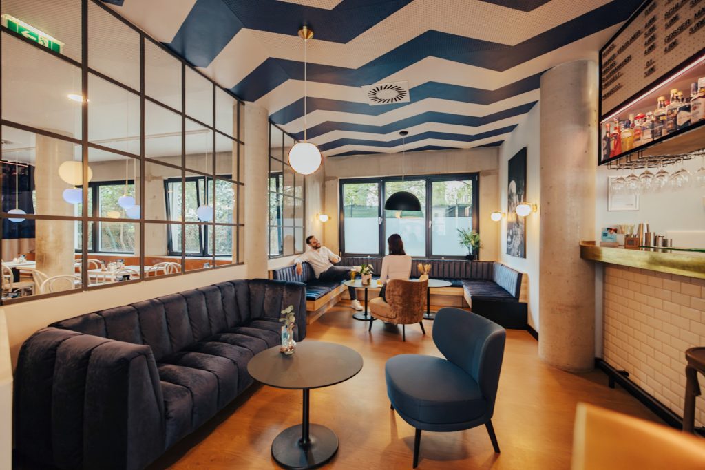 Hotellounge mit blauen Samtsofas, gestreifter Decke und Bar im MEININGER Wien