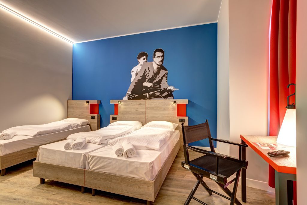 Sauberes Hotelzimmer mit Doppel- und Einzelbett, blauer Akzentwand und einem Vintage-Vespa-Wandbild