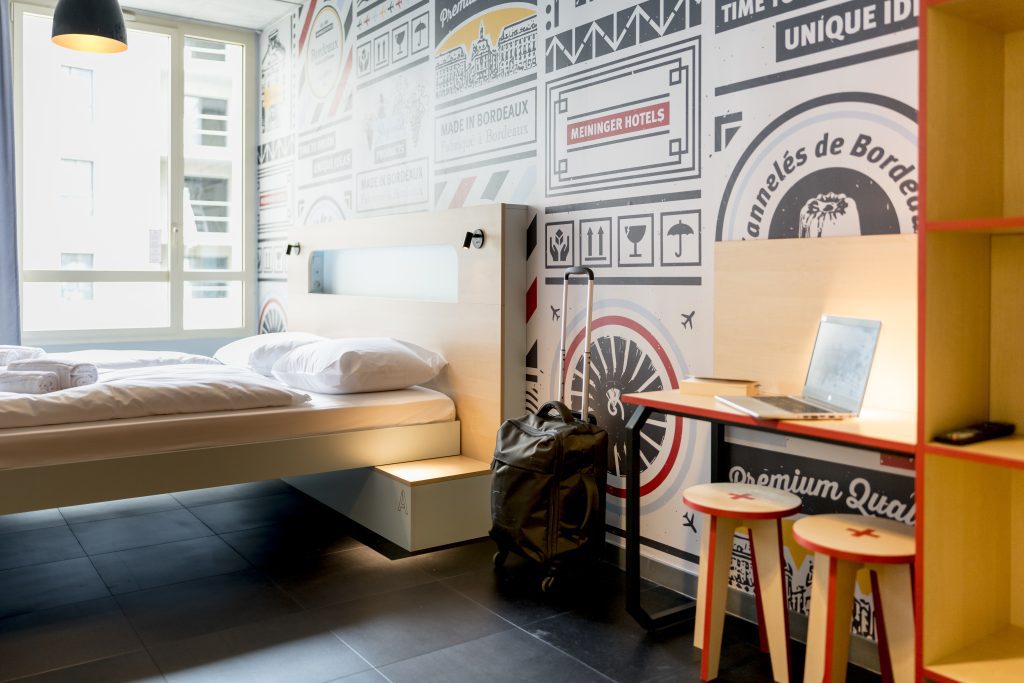 Bed, luggage, desk with laptop by a Bordeaux-themed wall