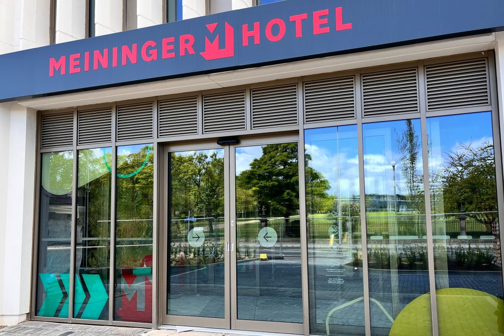 Glasschiebetüren unter einem dunkelblauen Schild. Bäume und eine Straße spiegeln sich im Glas.