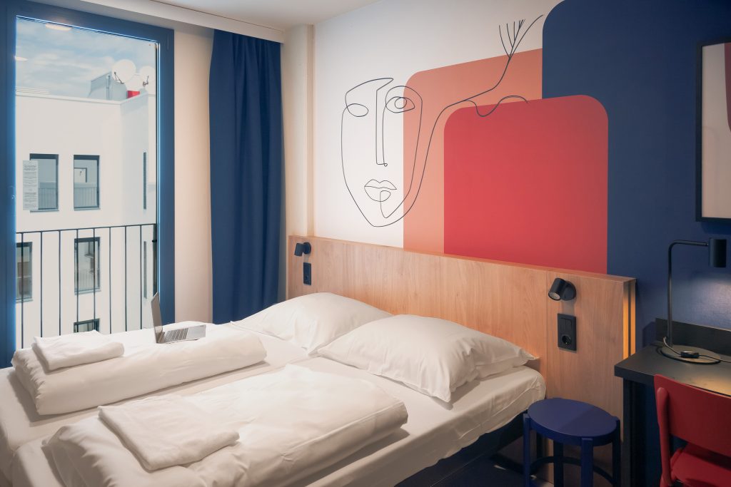 Hotel room with a bed, wall art mural and balcony window in blue tones