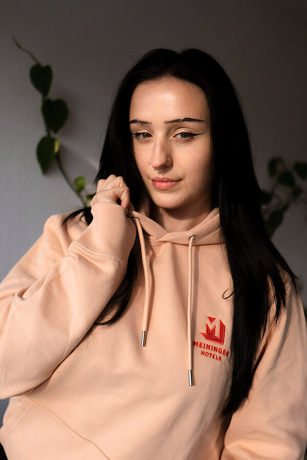 A portrait of Emmo with a MEININGER Hotels branded peach hoodie
